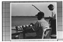 Speed Boats.  Two men sitting at a desk on a dock by water.